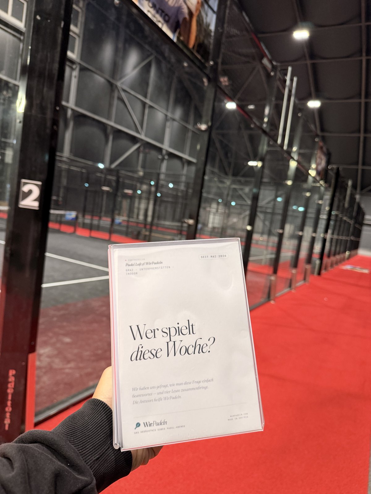 WirPadeln-Aufsteller in der Hand vor den Padel-Courts vom Padel Loft Graz